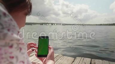 女孩躺在河边的木墩上，看着绿色的智能手机屏幕，用手指放大它